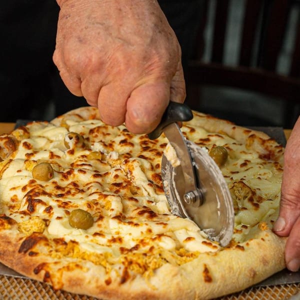 Hands cutting a cheesy pizza topped with olives and chicken using a pizza cutter.